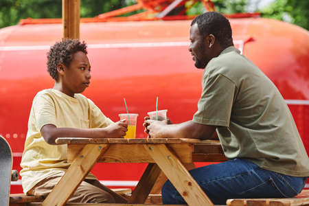 Dad talking to his son sitting at table during their rest at outdoor cafe with cold juiceの写真素材