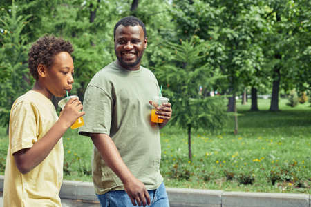 Happy African dad drinking cold juice and having a stroll with his son in the park during summer vacationの写真素材