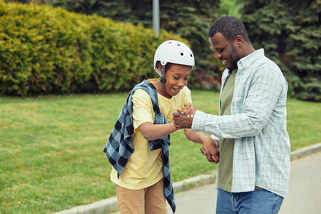 African boy in helmet holding hands with his dad while learning to ride on skateboard outdoorsの写真素材