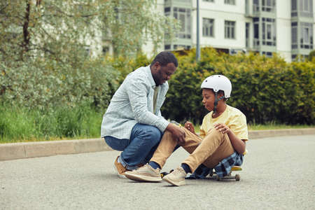 African dad putting protection on knees to his son before skateboarding outdoorsの写真素材