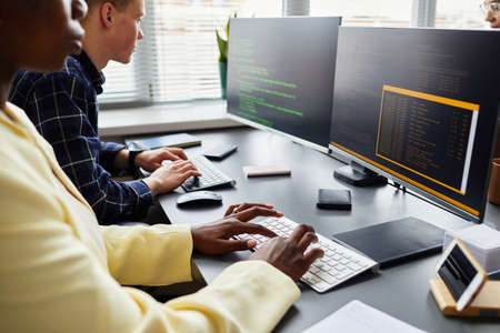 Close-up of colleagues sitting at table in front of monitors and typing on keyboard, they working...