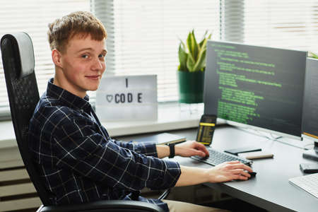 Portrait of young programmer looking at camera while sitting at his workplace with computer monitor...
