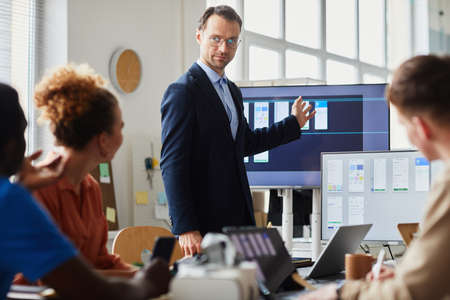Speaker in suit pointing at monitor with computer interface during meeting with his colleagues at officeの写真素材