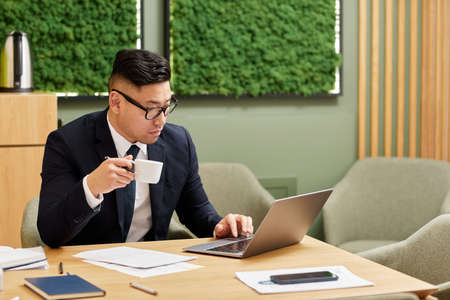 Portrait of successful Asian business expert working with laptop in office lounge and drinking...
