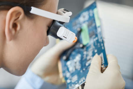Close up of young woman wearing magnifying visor inspecting computer parts in engineering lab, copy...