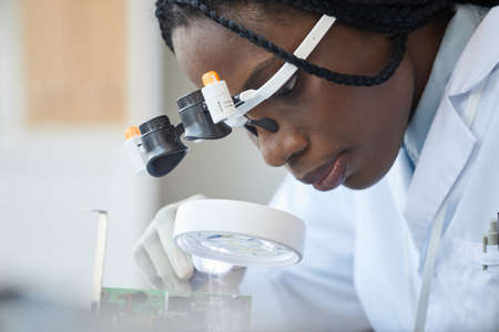 Close up portrait of black female engineer inspecting computer parts with magnifier at quality control lineの写真素材