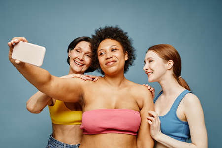 Waist up portrait of carefree young women taking selfie photo against pastel blue background and...