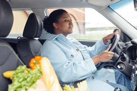 Side view portrait of young black woman driving car with grocery bag in foreground