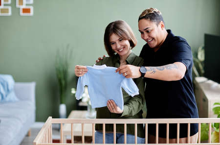 Waist up portrait of happy lesbian couple expecting baby and preparing crib with baby clothes at home, same sex family, copy spaceの写真素材