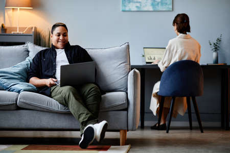 Portrait of modern multiethnic woman relaxing on couch at home and using laptop, copy spaceの写真素材