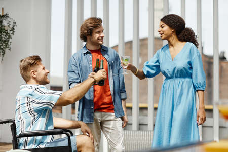 Diverse group of friends with person in wheelchair clinking glasses while enjoying party at outdoor terrace lit by sunlightの写真素材