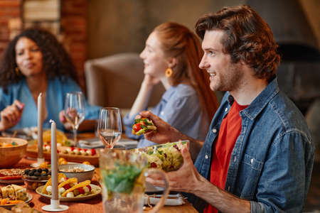 Side view portrait of young man talking to friends at table during dinner party in cozy setting, copy spaceの写真素材