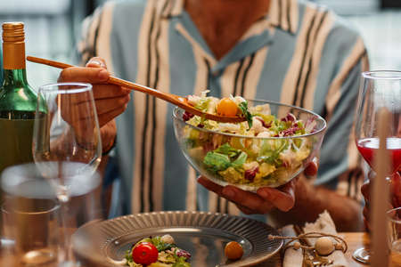 Close up of young man holding salad bowl while enjoying dinner party in cozy setting, copy spaceの写真素材
