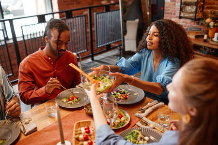 High angle portrait of diverse young people enjoying dinner party at table in cozy setting and handing salad bowlの写真素材