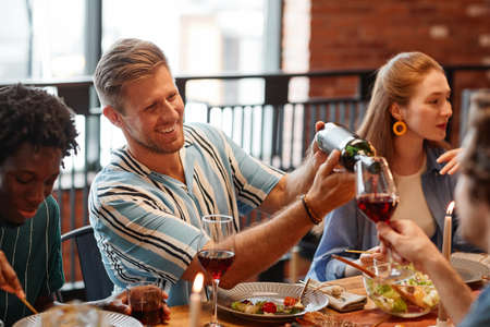 Portrait of smiling man pouring wine into glass while enjoying dinner party with friends in cozy...