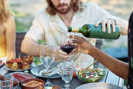 Close up of couple pouring red wine into glass while enjoying dinner party with friends outdoors in Summerの写真素材