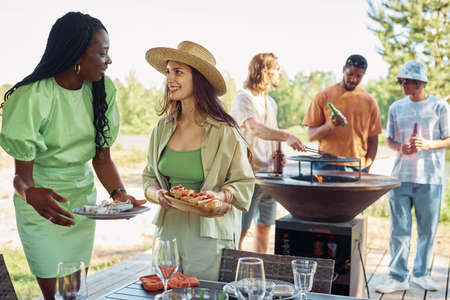 Diverse group of young people enjoying barbeque party outdoors in Sumer, focus on two smiling women serving food, copy spaceの写真素材