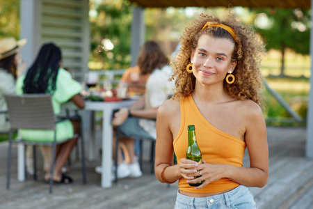 Waist up portrait of curly haired young woman smiling at camera and holding drink during outdoor party with friends in Summer, copy spaceの写真素材