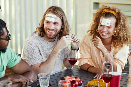Portrait of smiling man with sticker note on head playing Guess who game with group of friends at...