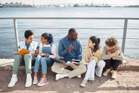 Full length portrait of smiling male teacher with diverse group of children enjoying outdoor class by water in sunlightの写真素材