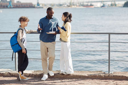 Full length portrait of black male teacher talking to teenage students outdoors while standing by water, copy spaceの写真素材