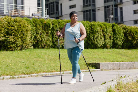Full length portrait of smiling overweight woman walking outdoors with nordic poles and enjoying cardio workout in sunlightの写真素材