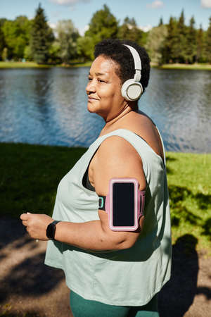 Vertical portrait of mature black woman running outdoors and wearing headphones enjoying workoutの写真素材