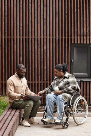 Vertical full length shot of young woman in wheelchair talking to friend or colleague outdoors in...