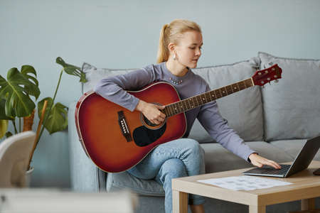 Minimal portrait of blonde young woman playing guitar at home and composing music while sitting on couch, copy space