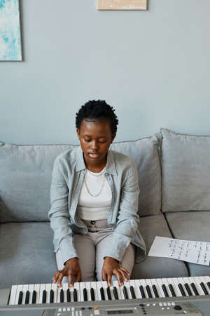 Vertical portrait of young black woman playing synthesizer at home and composing music in minimal...