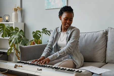Portrait of young black woman playing synthesizer at home and composing music in cozy home setting, copy spaceの写真素材