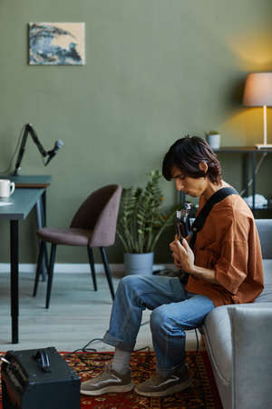 Vertical side view portrait of young man playing electric guitar in home music studioの写真素材