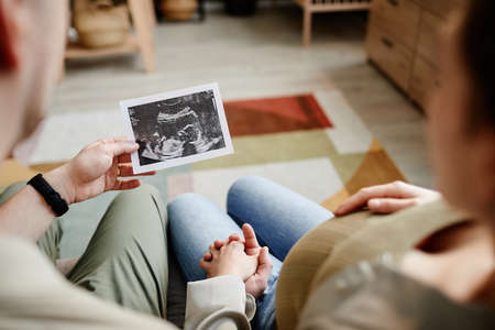 Rear view of affectionate young couple looking at ultrasound picture of their baby while sitting on...