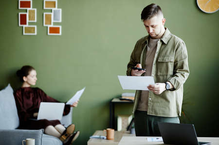 Young man taking photo of document on his mobile phone during his work at home with woman working in...