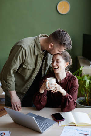 Young man kissing his girlfriend and supporting her while she sitting at table drinking coffee and working online at homeの写真素材