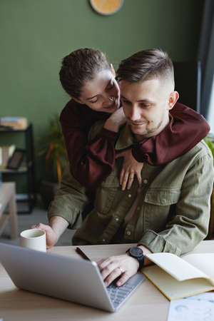 Young smiling woman embracing her boyfriend while he doing his online work on laptop at homeの写真素材
