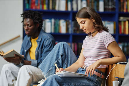 Vibrant side view portrait of young woman doing homework in college library and writing in notebook