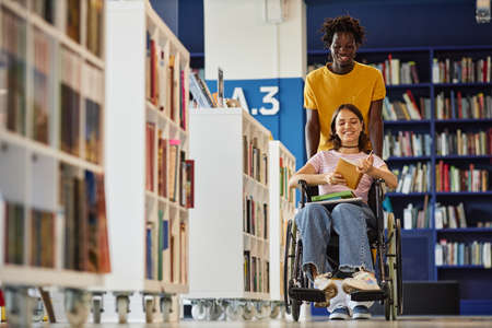 Vibrant full length shot of black young man assisting female student with disability in library and...