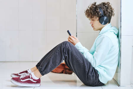 Side view portrait of depressed teen schoolboy sitting on floor and using smartphone with headphonesの写真素材