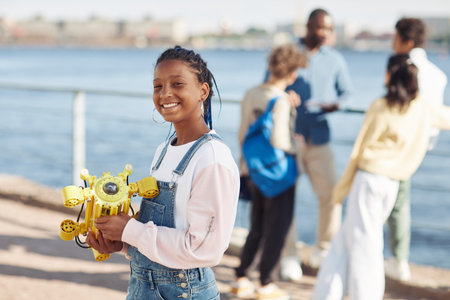 Waist up portrait of black teenage girl holding robot model and smiling at camera during outdoor engineering class with kids in background, copy spaceの写真素材