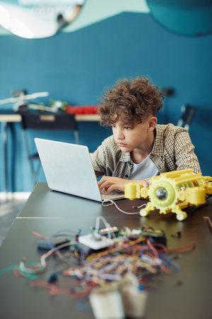 Vertical portrait of young curly haired boy using laptop in school during engineering class and programming robotの写真素材