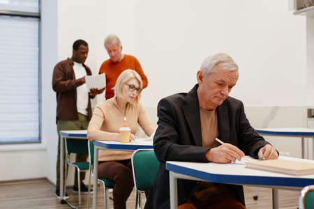 Senior people sitting at desks in a row and writing test at classroom with teacher in backgroundの写真素材