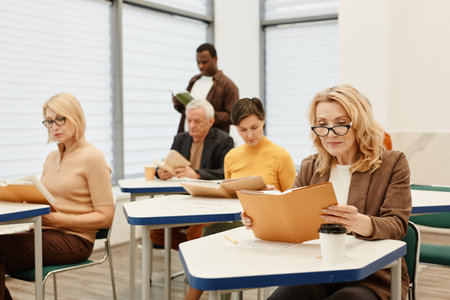 Mature woman in eyeglasses sitting at her desk and reading a book during lecture at class with teacher and students in backgroundの写真素材