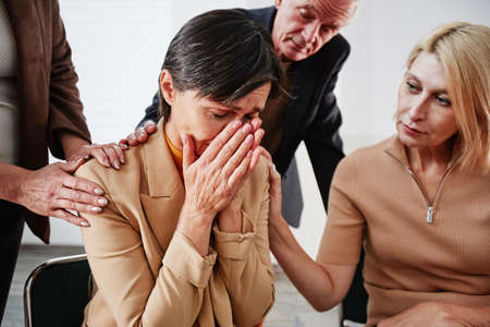 Mature woman with short hair sitting on chair and crying while people supporting her during psychotherapy sessionの写真素材