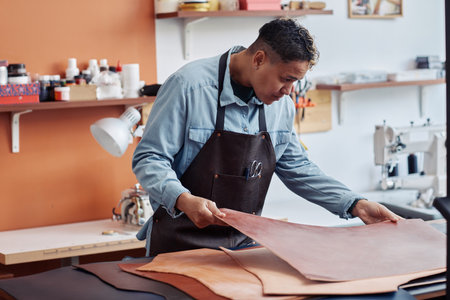 Side view portrait of young artisan inspecting leather in workshop and creating handmade pieces,...