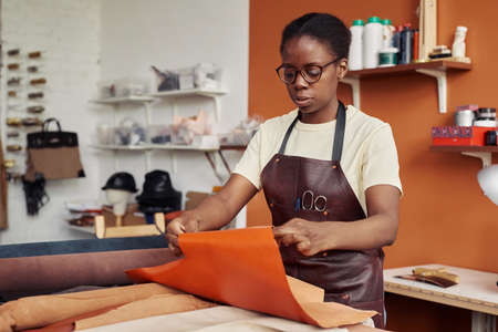 Portrait of black female artisan creating handmade leather pieces in shop and working with genuine...