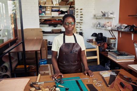Waist up portrait of young female artisan standing in leatherworking shop and smiling at camera...