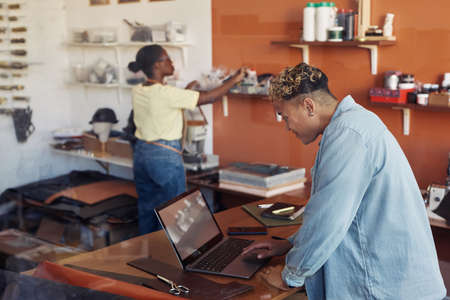 Side view portrait of female small business owner using computer in leatherworking workshop, copy spaceの写真素材
