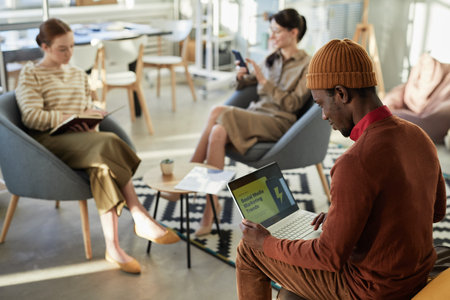 Side view portrait of young black man typing at laptop with marketing statistics on screen in...