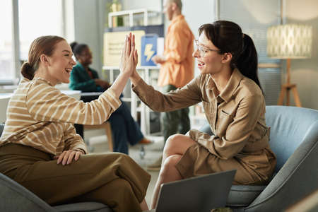 Side view portrait of two young women high five in office celebrating success in businessの写真素材